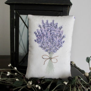Puede incluir: Una almohada blanca con una ilustraci&oacute;n de un ramo de lavanda. La almohada est&aacute; atada con una cinta marr&oacute;n y tiene un fondo marr&oacute;n claro.