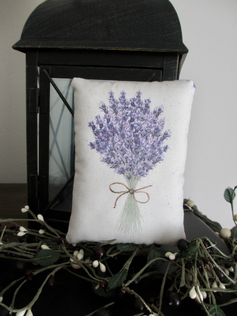 Puede incluir: Una almohada blanca con una ilustraci&oacute;n de un ramo de lavanda. La almohada est&aacute; atada con una cinta marr&oacute;n y tiene un fondo marr&oacute;n claro.