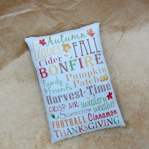 May include: White pillow with a brown border and a colorful design featuring the words "Autumn leaves Fall Cider Bonfire Family & Friends Pumpkin Patch Harvest Time Crisp Air sweater Scarecrow weather Football Cinnamon Thanksgiving".