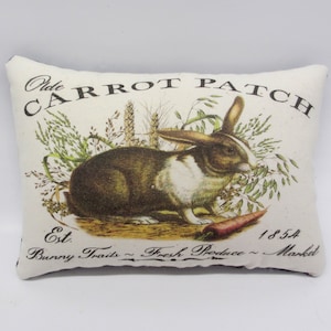 May include: A white pillow with a brown rabbit and green foliage. The pillow has black text that reads "Olde Carrot Patch Est. Bunny Traits - Fresh Produce - Market 1854".