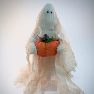 May include: A decorative ghost figurine draped in off-white, semi-sheer cheesecloth fabric. The ghost features two small black bead eyes and holds a round, orange fabric pumpkin with a brown stem and green moss accents in its center.