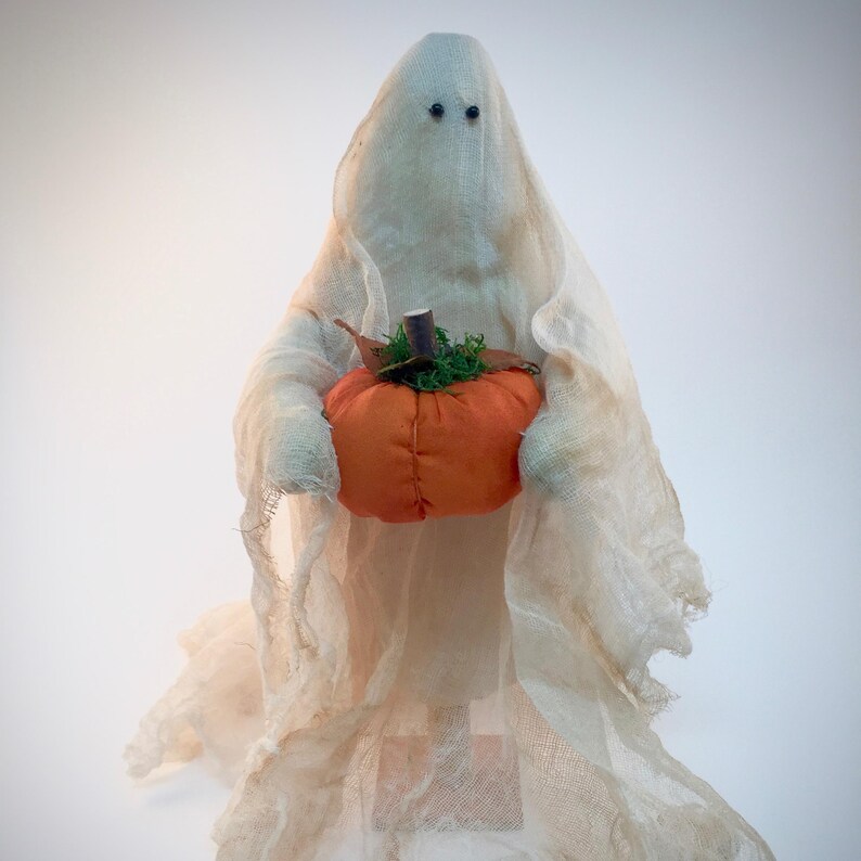 May include: A decorative ghost figurine draped in off-white, semi-sheer cheesecloth fabric. The ghost features two small black bead eyes and holds a round, orange fabric pumpkin with a brown stem and green moss accents in its center.