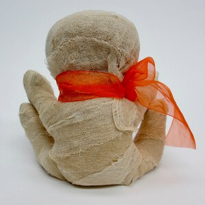 May include: A white fabric mummy doll with a bright orange ribbon tied around its head.
