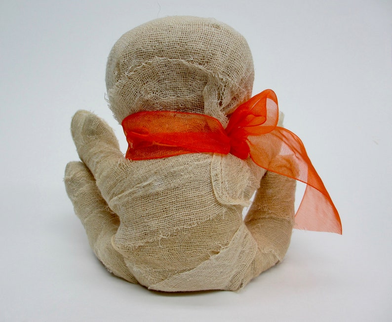 May include: A white fabric mummy doll with a bright orange ribbon tied around its head.