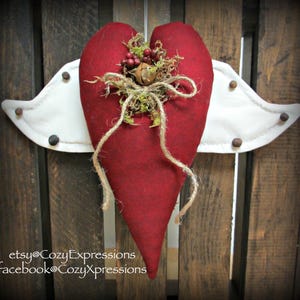 May include: A fabric heart ornament in deep red with attached white fabric wings. The heart is decorated with green moss, small red berries, a metal bell, and a jute twine bow. Dark brown circular accents are stitched onto the white wings.