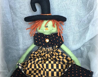 May include: A fabric doll featuring a light green face, orange yarn hair, and a black pointed hat. The doll wears a black bodice with orange polka dots and a skirt with a black and yellow checkered pattern, finished with a black polka dot hem.