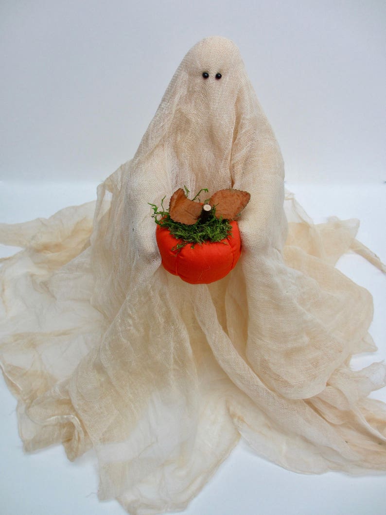 May include: A decorative ghost figurine draped in off-white, semi-sheer cheesecloth fabric. The ghost has two small black bead eyes and holds a bright orange fabric pumpkin topped with green moss and two brown felt leaves in its center.