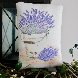 Puede incluir: Una almohada blanca con una imagen impresa de un cubo de flores de lavanda. El cubo es blanco con un asa marr&oacute;n. La almohada est&aacute; sobre una superficie marr&oacute;n con hojas verdes y bayas blancas.