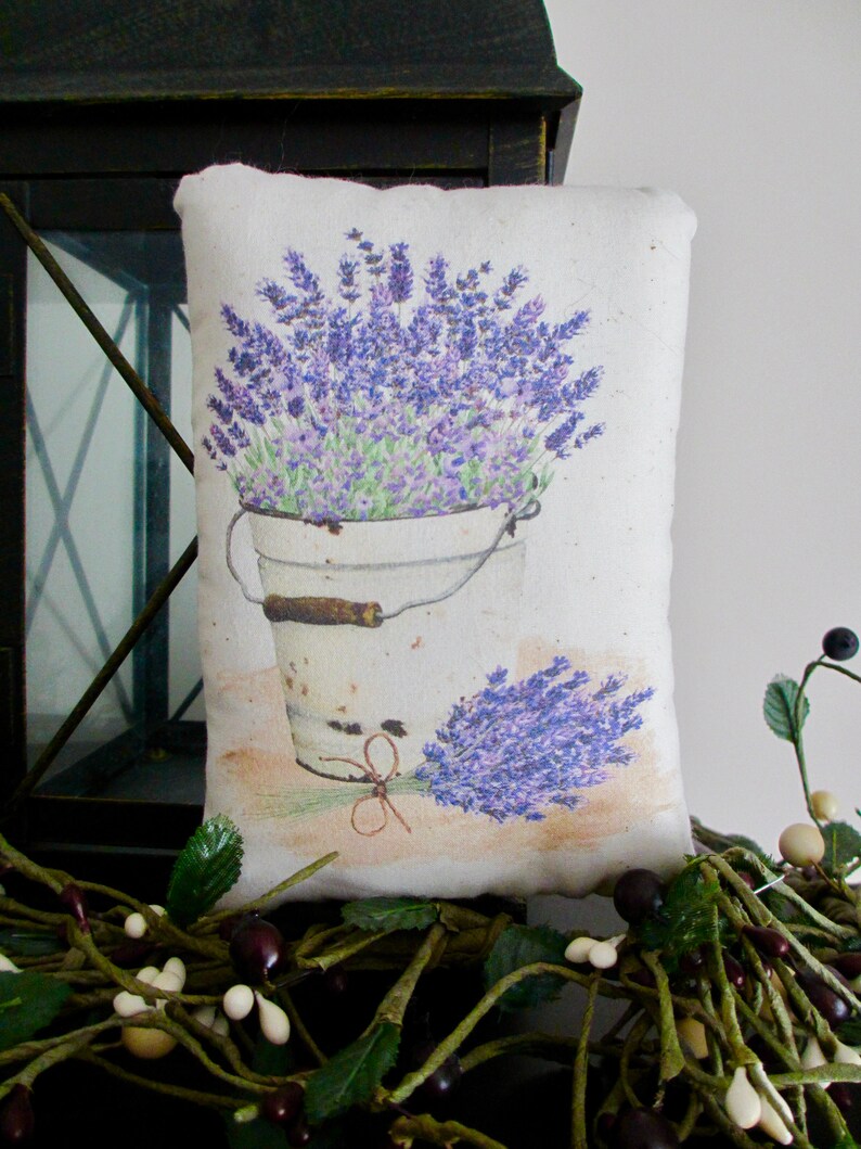 Puede incluir: Una almohada blanca con una imagen impresa de un cubo de flores de lavanda. El cubo es blanco con un asa marr&oacute;n. La almohada est&aacute; sobre una superficie marr&oacute;n con hojas verdes y bayas blancas.