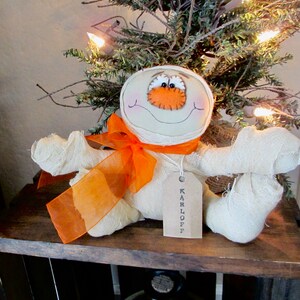 May include: A white fabric mummy doll with an orange ribbon and a tag that says "KARLOFF". The doll has a stitched-on orange nose and black eyes.