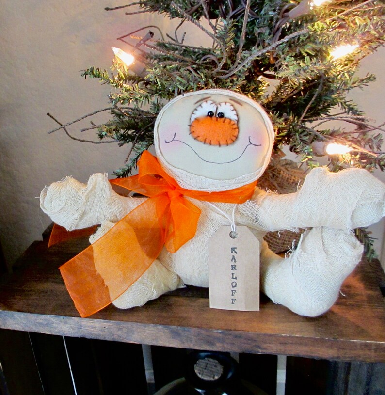 May include: A white fabric mummy doll with an orange ribbon and a tag that says "KARLOFF". The doll has a stitched-on orange nose and black eyes.