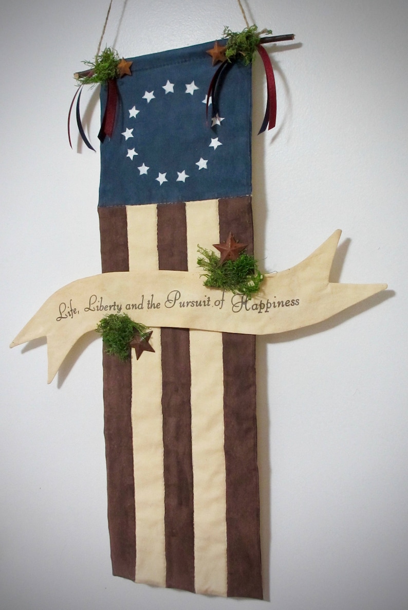 May include: A decorative wall hanging featuring a fabric American flag with 13 stripes and a blue canton with 13 white stars. A wooden banner with the text "Life, Liberty and the Pursuit of Happiness" is attached to the bottom of the flag. The flag is decorated with green moss and small, brown, star-shaped embellishments.