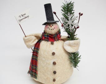 May include: A fabric snowman figurine wearing a black top hat and a red, green, and yellow plaid scarf. The snowman has four dark buttons on its body, holds a green pine branch with a silver snowflake, and carries a flag reading I Love Snowdays.