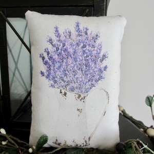 Puede incluir: Una almohada blanca con un ramo de lavanda en una jarra impresa en ella. La almohada est&aacute; sentada sobre una superficie marr&oacute;n con enredaderas verdes.