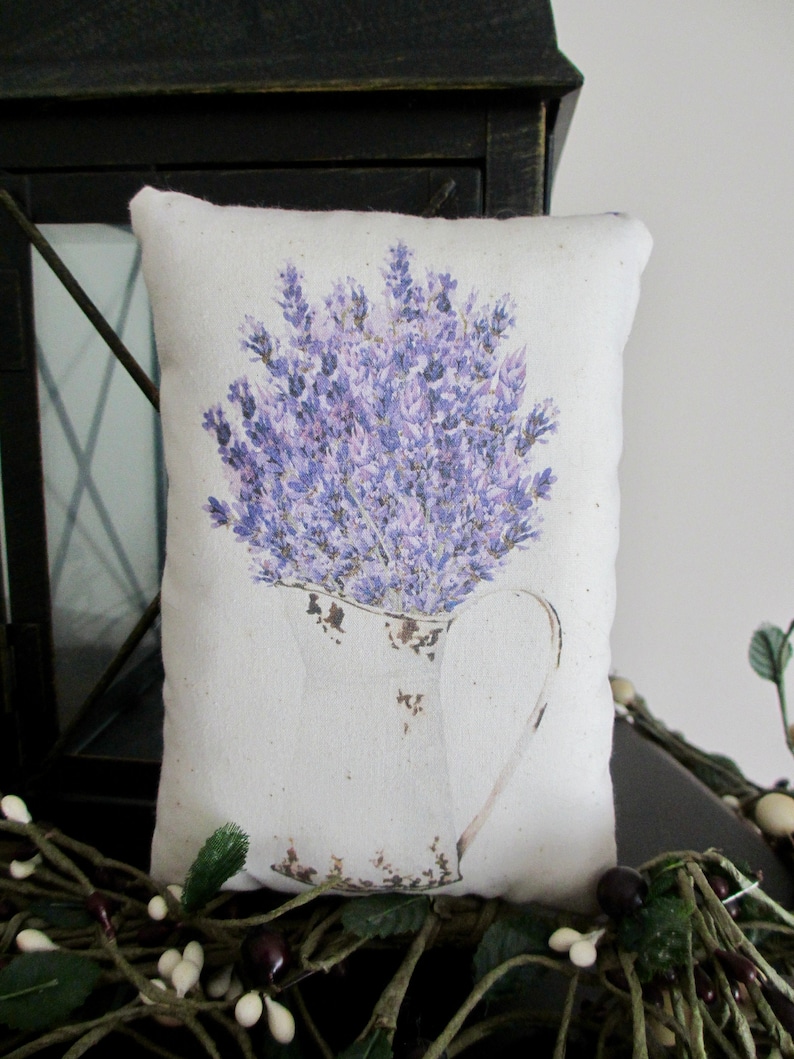Puede incluir: Una almohada blanca con un ramo de lavanda en una jarra impresa en ella. La almohada est&aacute; sentada sobre una superficie marr&oacute;n con enredaderas verdes.