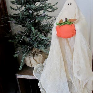May include: A fabric ghost figure draped in off-white cheesecloth with two small black bead eyes, holding a small, round, orange fabric pumpkin topped with green faux moss and brown fabric leaves, positioned next to a small artificial pine tree.