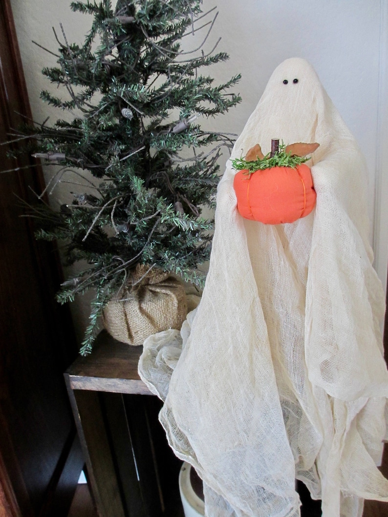 May include: A fabric ghost figure draped in off-white cheesecloth with two small black bead eyes, holding a small, round, orange fabric pumpkin topped with green faux moss and brown fabric leaves, positioned next to a small artificial pine tree.
