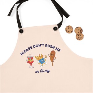 Op de afbeelding: Een beige keukenschort met zwarte banden en twee zwarte knopen. Het schort heeft een blauwe en witte afbeelding van een cartoon-frisdrank, frietjes en een corn dog met de tekst "PLEASE DON'T RUSH ME or I'll cry".