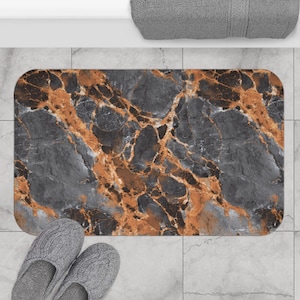 May include: Rectangular bath mat with a marble design in shades of gray and orange. The mat has rounded corners and is placed on a tiled floor. A pair of gray knitted slippers are in the foreground.