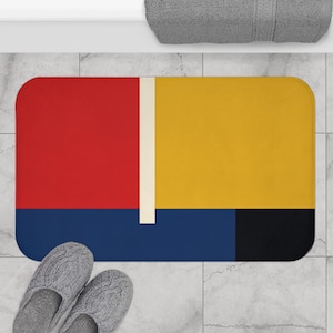 May include: A bath mat with a geometric design featuring red, yellow, blue, and black squares. The mat has a white stripe running down the center.