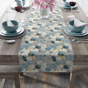 May include: A table runner with a blue, gray, and white bubble pattern, accented with gold lines. The runner is placed on a wooden table set for a meal, with plates, bowls, and wine glasses.