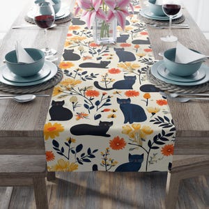 May include: A cream-colored table runner with a repeating pattern of black and blue cats, yellow and orange flowers, and green leaves.