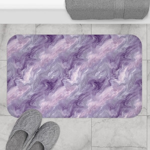 May include: A rectangular bath mat with a swirling pattern in shades of purple, lavender, and white. The mat is placed on a tiled floor, with a pair of gray slippers at the bottom. A rolled gray towel is visible in the background.