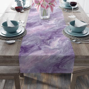 May include: A purple and white abstract patterned table runner. The runner is draped over a wooden table set with plates, bowls, silverware, and wine glasses. A vase of pink flowers sits in the center.