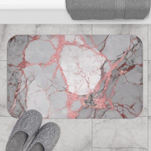 May include: A rectangular bath mat with a marble design. The mat features a gray and white background with rose gold veining. A pair of gray knit slippers are at the bottom of the mat.