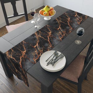 May include: A black dining table with a decorative runner featuring a gold marble design. The table is set with white plates, silverware, wine glasses, and a bowl of apples. The chairs are black with brown seat cushions.