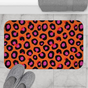 May include: Orange rectangular bath mat with a bold leopard print pattern in black and pink. The mat is placed on a tiled floor, with a pair of gray slippers and a gray towel visible.