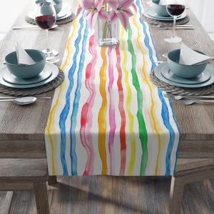 May include: A colorful table runner with a rainbow stripe pattern. The stripes are painted in a watercolor style and are slightly wavy. The runner is on a wooden table with place settings.