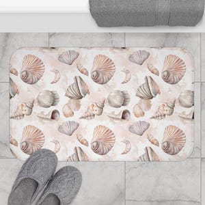 May include: A pink and white bath mat with a seashell pattern. The mat features various seashells in shades of pink, white, and brown, with a watercolor-like effect.