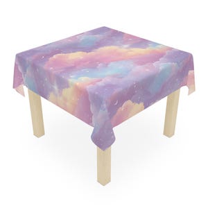 May include: A square tablecloth with a dreamy, pastel cloud and starry night sky design. The colors include shades of purple, pink, yellow, and blue. The tablecloth is draped over a small, light-colored wooden table.