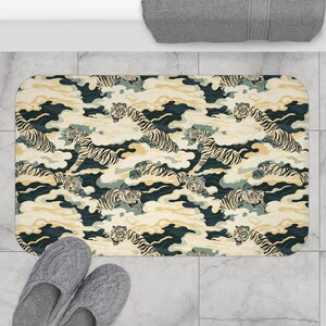 Puede incluir: Alfombra de baño rectangular con un patrón repetido de tigres entre nubes estilizadas. El diseño incorpora tonos crema, azul marino y dorado. La alfombra está colocada sobre un suelo embaldosado, con un par de zapatillas grises visibles.
