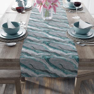 May include: A gray and teal marble-patterned table runner is displayed on a wooden table, set for a meal. The table is set with teal bowls, plates, and wine glasses. A vase of pink lilies sits in the center.