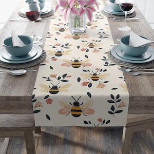 May include: A beige table runner featuring a repeating pattern of bees, flowers and leaves. The bees are yellow and black with brown wings. The flowers are pink and orange. The leaves are green.