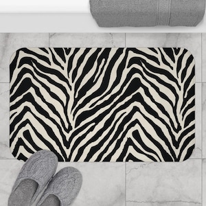 May include: A rectangular bath mat with a black and white zebra stripe pattern. The mat is placed on a tiled floor, with a pair of gray slippers and a gray towel visible.