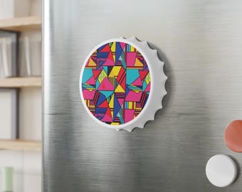 May include: A white bottle cap magnet with a colorful geometric design. The design features triangles and lines in shades of pink, yellow, blue, and black. The magnet is attached to a stainless steel refrigerator.