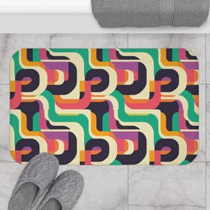 May include: A bath mat with a geometric pattern of curved lines in shades of green, orange, pink, purple, and black on a cream background.