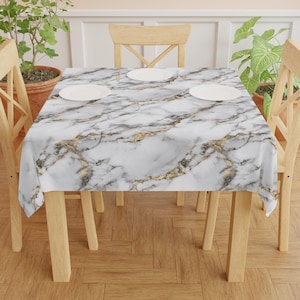 Pode incluir: Toalha de mesa de mármore branco com veios dourados, cobrindo uma mesa de madeira com quatro cadeiras. A toalha de mesa é quadrada e está colocada plana sobre a mesa.