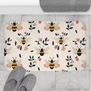 May include: A white bath mat with a repeating pattern of bees and flowers. The bees are yellow and black with brown wings. The flowers are pink and white.
