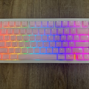 May include: A white mechanical keyboard with pink keycaps and a rainbow LED backlight. The keyboard has a volume knob and a small display screen. The keys are arranged in a standard layout.