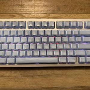 Puede incluir: Un teclado mecánico plateado con teclas azul claro y una pequeña figura blanca en la parte superior izquierda. El teclado tiene una perilla de volumen y una pequeña pantalla. Las teclas están retroiluminadas en púrpura.