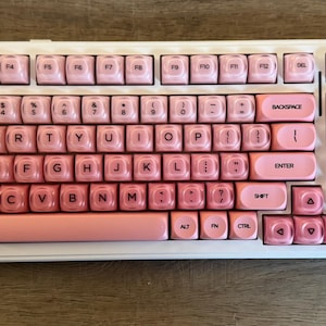 May include: A white and pink mechanical keyboard with pink keycaps. The keycaps have a gradient effect, transitioning from light pink to a darker shade. The keyboard has a standard QWERTY layout with a numeric keypad.