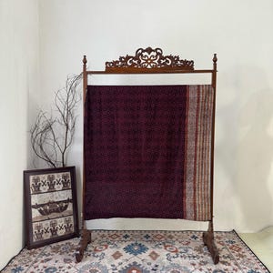 Op de afbeelding: Een donker bordeauxrood textiel met een geometrisch patroon wordt getoond op een houten rek. Het rek heeft een sierlijke bovenkant en staat voor een witte muur. Een ingelijst kunstwerk en een patroonkleed zijn ook zichtbaar.
