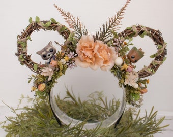 May include: A brown twig headband with a peach-colored flower, green leaves, and small figurines of a raccoon and a tree-like creature.