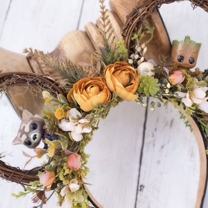 May include: A pair of brown twig Mickey Mouse ears with a brown headband. The ears are decorated with faux flowers, including orange roses, white flowers, and green leaves. A small brown Groot figurine is nestled in the right ear, and a small brown Rocket Raccoon figurine is nestled in the left ear.