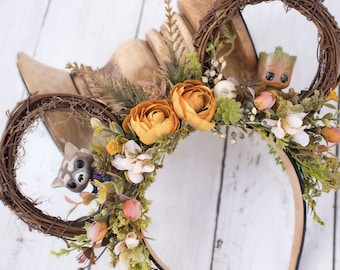 May include: A pair of brown twig Mickey Mouse ears with a brown headband. The ears are decorated with faux flowers, including orange roses, white flowers, and green leaves. A small brown Groot figurine is nestled in the right ear, and a small brown Rocket Raccoon figurine is nestled in the left ear.