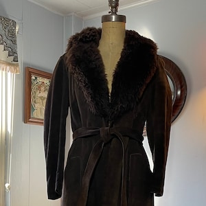 May include: A brown suede coat with a faux fur collar and a tie belt. The coat is on a mannequin and is displayed against a light blue wall.